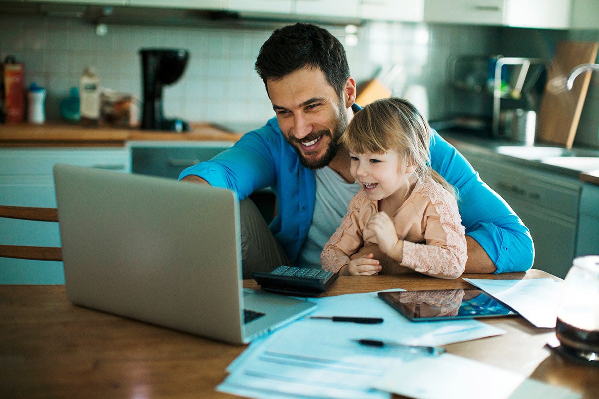 a man and his daughter at a kitchen table looking at computer with papers on the table